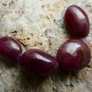 May include: Four polished, oval-shaped, deep red gemstones with a glossy finish are displayed on a textured, light brown stone surface. The gemstones vary slightly in size and shape, with visible internal patterns.