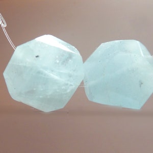 May include: Two faceted, light blue gemstone beads strung on a thin, clear cord. The beads have a hexagonal shape with multiple facets, giving them a sparkling appearance. The background is a soft, neutral color. The text "No 2" is in the lower right corner.
