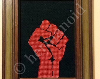 May include: A framed cross-stitch artwork depicting a red fist raised in the air against a black background.