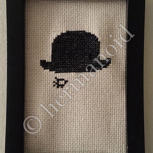 May include: A black and white cross-stitch of a bowler hat in a black frame.
