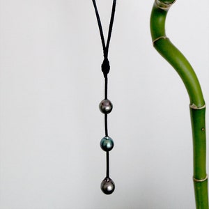 May include: A black cord necklace with three dark green and blue pearls hanging from it. The necklace is hanging from a green bamboo plant.