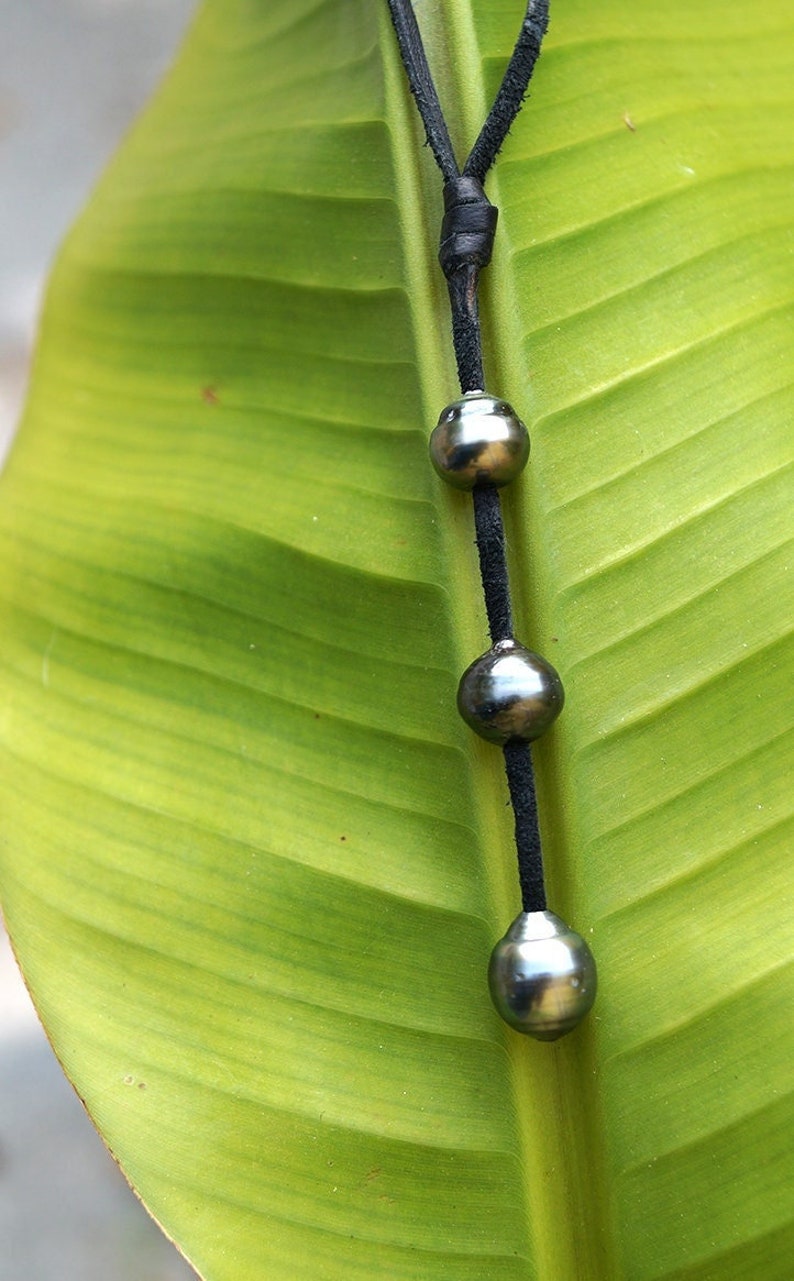 May include: A black leather cord necklace with three dark gray round pearls. The necklace is laying on a green leaf.