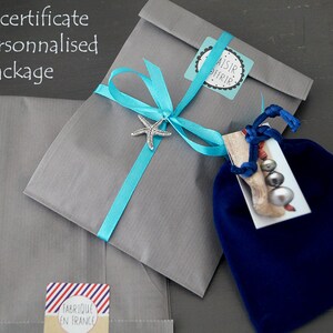 May include: A gray gift bag with a blue ribbon and a starfish charm. The bag is tied with a blue ribbon and has a small tag that says "aisir offrir". The bag is sitting on a gray surface. The bag is also labeled "fabrique en france".