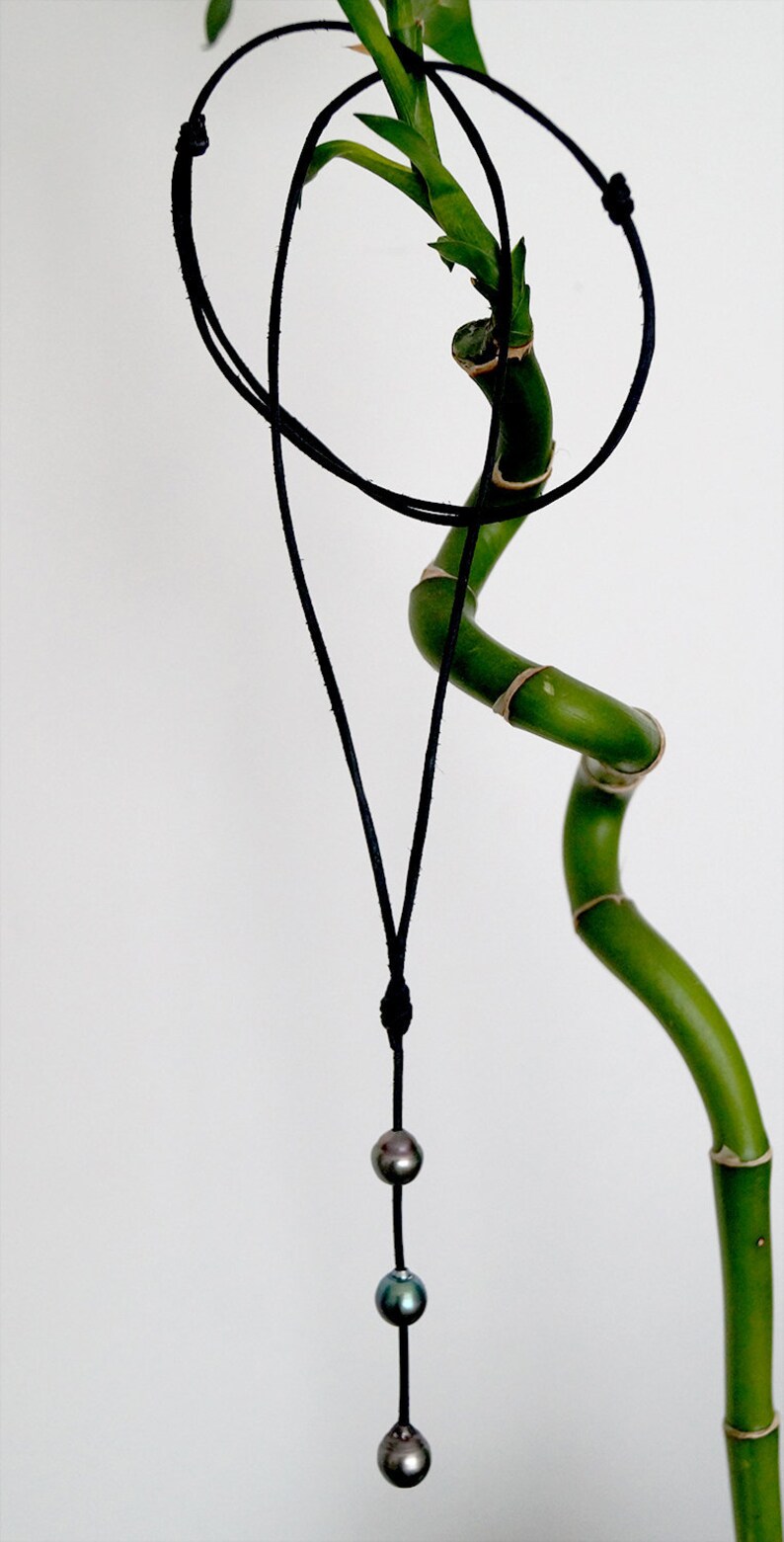 May include: A black cord necklace with three dark green and blue pearls hanging from it. The necklace is hanging from a green bamboo plant.