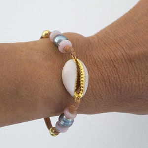 Peut inclure: Un bracelet orné d'un coquillage blanc avec une bordure dorée. Le bracelet est composé de perles roses, grises et marron, ainsi que de détails dorés. Le bracelet est porté au poignet.
