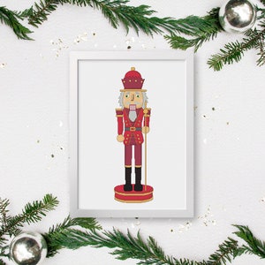 May include: A framed print featuring a cross-stitch design of a traditional Christmas nutcracker soldier. The nutcracker is depicted in shades of red, gold, and black, with a detailed uniform and a regal hat. The artwork is set against a white background.
