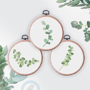 May include: Three embroidery hoops with cross-stitch patterns of eucalyptus leaves in shades of green. The hoops are made of wood and have a natural finish.