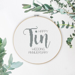 May include: A circular cross-stitch embroidery hoop with the words "Happy Tin Wedding Anniversary" in grey. The word "Tin" is in a large, decorative font. The hoop is surrounded by eucalyptus leaves.