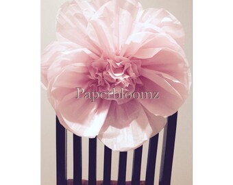 May include: A large, pale pink paper flower, possibly tissue paper, rests on a dark wooden chair. The flower has multiple layers of petals, with the text "Paperbloomz" visible. The chair's back features vertical slats.
