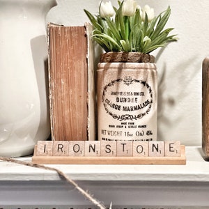 May include: A vintage-style display featuring a white ceramic vase, an antique book, and a Dundee Orange Marmalade jar with white tulips. The word "IRONSTONE" is spelled out with wooden letter tiles. The jar is labeled "JAMES KEILLER & SON LTD. DUNDEE ORANGE MARMALADE".