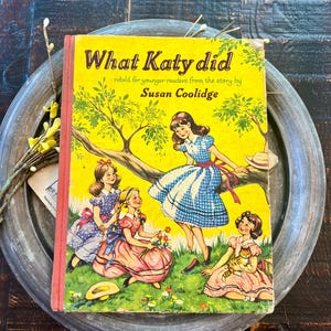 May include: A vintage children's book titled "What Katy Did" with a yellow cover and illustrations of girls in dresses. The cover features the title in large, bold letters and the author's name. The book is set on a metal tray.