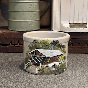 May include: A hand-painted ceramic jar featuring a scene of a covered bridge in a woodland setting. The bridge is painted in shades of brown, black, and green, with white accents. The jar is a light beige colour.