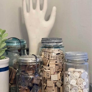 May include: A collection of vintage jars filled with various craft supplies. The jars contain wooden letter tiles, buttons, and other small items. A white ceramic hand sculpture is in the background.