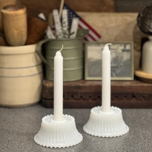 May include: Two white taper candles standing in matching white milk glass candle holders. Each holder features a flared base with a vertical ribbed pattern and a beaded edge along the bottom rim. The candles are slender, cylindrical, and plain.