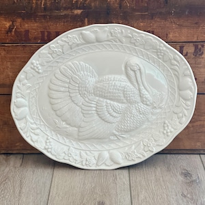 May include: An oval-shaped cream-colored ceramic serving platter featuring a raised, embossed turkey design in the center. The rim is decorated with a detailed relief pattern of fruits, leaves, and flowers, framed by a woven basket-weave border.
