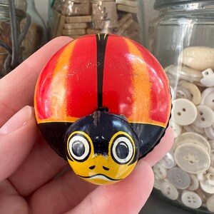 May include: A red, yellow, and black wind-up toy ladybug with a black and yellow face. The ladybug has large, round eyes and a small, yellow mouth.