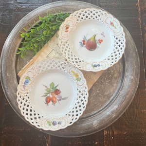 May include: Two white porcelain plates with a lattice design and floral accents. One plate features a pear and a fig, while the other plate features a branch of red berries.