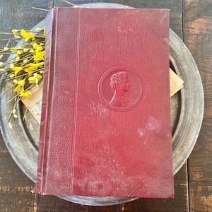May include: A vintage, red hardcover book with a textured cover and a circular embossed portrait. The book is resting on a metal tray, with yellow flowers and aged paper visible. The book's spine is visible.