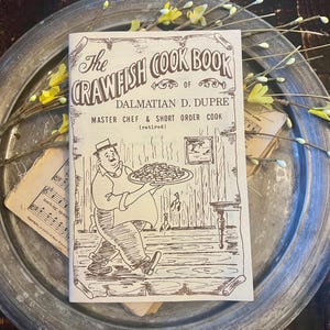 May include: A softcover book titled The Crawfish Cook Book of Dalmatian D. Dupre, Master Chef & Short Order Cook (retired). The cover features a brown line drawing of a chef carrying a platter of food, framed by a scroll-like border design.