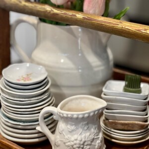 May include: A white ceramic pitcher with a grape design and a stack of small plates on a wooden tray. Pink tulips are in a large white pitcher in the background. A small white creamer is in the foreground.