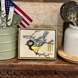 May include: A rectangular framed cross-stitch embroidery featuring a bird with a black head, white cheek, green wings, and a yellow breast perched on a brown branch. The artwork is displayed in a thin gold-toned metal frame with a light fabric background.