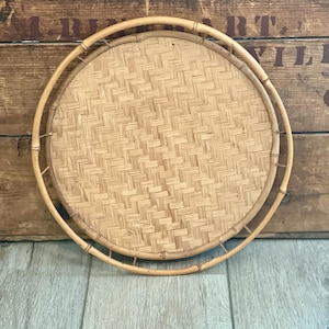 May include: A round, light brown woven bamboo tray featuring a flat, intricate basket-weave base and a raised, circular bamboo rim. The tray is constructed from natural, light-colored bamboo strips, showcasing a consistent, repeating pattern.