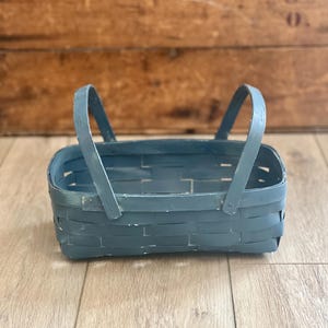 May include: A rectangular woven basket painted in a matte slate blue finish. The basket features a flat base, vertical sides constructed from interlaced strips, and two arched handles attached to the sides with small metal rivets.