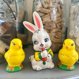 May include: A ceramic Easter bunny figurine with a white body, pink ears, and blue eyes, holding a carrot. Two yellow chick figurines with orange beaks and feet flank the bunny. All are on a green base.