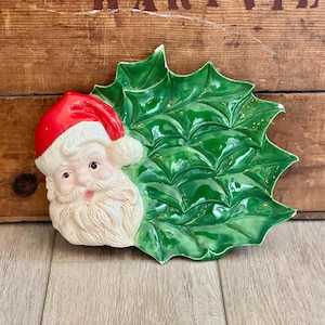 May include: A ceramic serving dish shaped like a large green holly leaf with a three-dimensional Santa Claus head attached to the left side. The Santa figure features a bright red hat, white beard, and pink cheeks, while the leaf has a glossy finish.