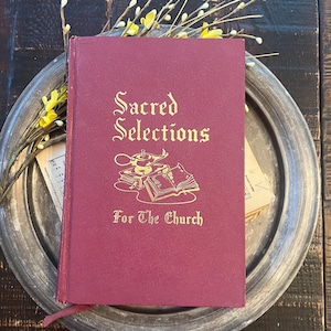 May include: A maroon book titled "Sacred Selections For The Church" with gold lettering and illustrations. The book is on a silver tray with yellow flowers and sprigs. The background is a dark wooden surface.