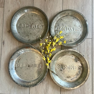 May include: Four vintage pie plates with the word "GARDNER" embossed on each. The plates are made of metal and have a rustic, weathered look. They are arranged in a square pattern on a wooden surface.
