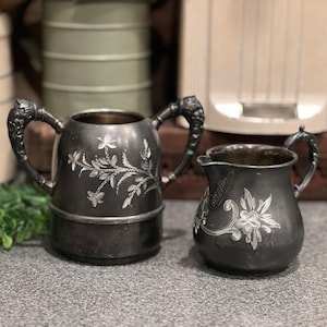 May include: A vintage silver creamer and sugar set with ornate floral designs. The creamer has two handles and the sugar bowl has a single handle.