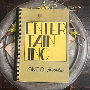 May include: A spiral-bound book with a muted olive green cover featuring the word ENTERTAINING in large black capital letters inside a rectangular border, with the text JANGO favorites written in a cursive script font below the main title.