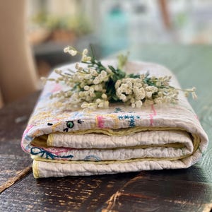 May include: Folded, cream-colored quilt with embroidered floral designs in pink, yellow, and blue. The quilt is edged with yellow fabric and topped with a small bouquet of white flowers. The quilt is folded neatly.
