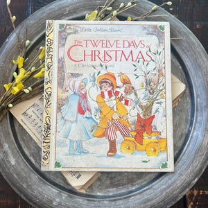 May include: A vintage "Little Golden Book" titled "The Twelve Days of Christmas." The cover shows illustrations of children in winter clothes, a bird, and a small cart. The book is on a silver-toned metal tray with floral decorations.