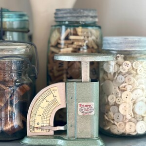 May include: A vintage sage green Triner Liberty No. 1 scale with a metal weighing platform and a curved dial showing measurements up to 1 lb. The scale features a red and black logo with a globe graphic and is positioned among glass jars.