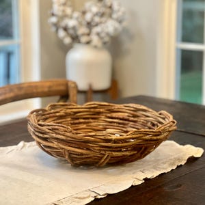 May include: A shallow, round basket constructed from thick, natural brown woven twigs. The bowl-shaped container features an open, rustic weave pattern with a textured, uneven rim, showcasing the organic, woody appearance of the intertwined branches.