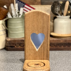 May include: A light brown wooden wall-mounted candle holder featuring a vertical rectangular back panel with a heart-shaped cutout in the center and a small circular base with a recessed center designed to hold a candle securely in place.