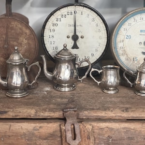 May include: A four-piece silver-toned metal tea set consisting of two teapots with lids, a small creamer pitcher, and a lidded sugar bowl. Each piece features a polished metallic finish, a pedestal base, and a curved handle, arranged in a row.