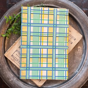 May include: A vintage book with a colorful plaid pattern in blue, green, and yellow. The book rests on top of sheet music and a metal tray, with a sprig of greenery. The plaid design is a classic, retro style.