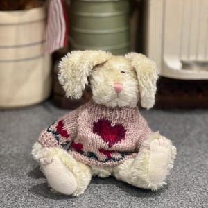 May include: A cream-coloured plush bunny rabbit with a pink nose. It wears a pink sweater with a red heart and green floral accents. The bunny is sitting on a grey surface.