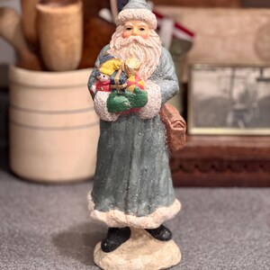 May include: A standing Santa Claus figurine wearing a long, textured, dusty blue coat with white faux fur trim, a matching hat, and black boots. The figure holds a collection of colorful wrapped gifts and wears a brown satchel over one shoulder.