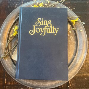 May include: A dark blue hardcover book with the words Sing Joyfully printed in gold lettering on the front cover. The book is positioned on a round, weathered metallic tray that features a decorative arrangement of thin branches with small yellow flowers.