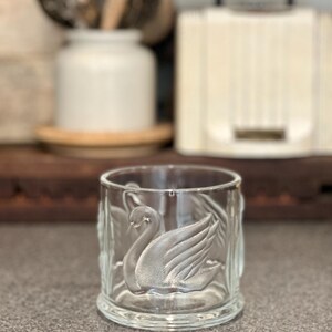 May include: A clear glass tumbler featuring a frosted, embossed swan design on its exterior. The cylindrical vessel has a smooth rim and a thick, sturdy base, with the swan motif appearing in relief against the transparent glass surface.