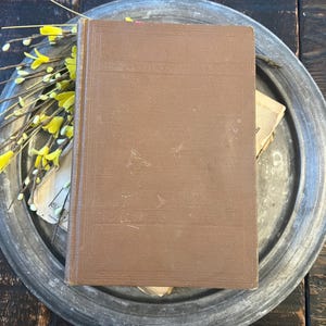 May include: A brown hardback book with a textured cover rests on a silver metal plate. Yellow floral sprigs are placed near the book. The plate sits on a dark wooden surface.