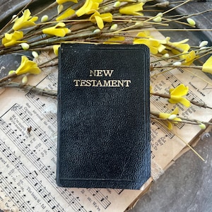 May include: A black, textured New Testament book with gold lettering on the cover. The book is placed over vintage sheet music and decorative yellow floral branches. The cover features the words NEW TESTAMENT in a clear, capitalized font.