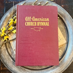 May include: A maroon hardcover book titled All-American Church Hymnal in gold lettering, featuring a debossed brick pattern and a gold outline of the United States map with stars on the front cover, resting on a round metallic tray with yellow flowers.