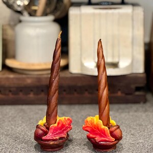 May include: Two brown spiral taper candles, each set in a decorative base featuring sculpted autumn leaves in shades of yellow, orange, and red, accented with small brown acorns at the base of the candle, creating a seasonal tabletop display.
