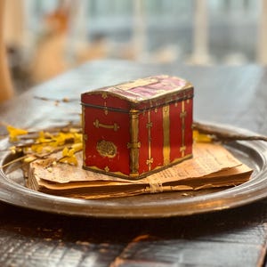May include: A red and gold decorative tin box shaped like a vintage suitcase. The box is sitting on a silver tray with a stack of paper underneath.