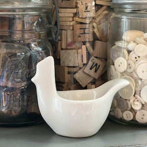 May include: A white ceramic bird-shaped egg cup sits on a gray surface in front of three glass jars filled with various items. The jars contain wooden letter tiles, buttons, and other small objects.